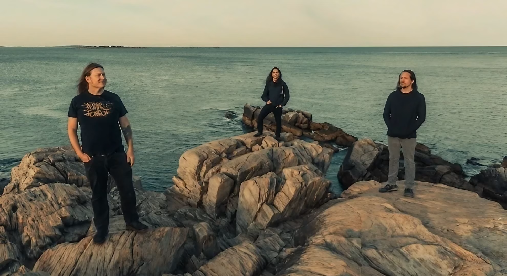 Shabti Send Servants on ‘Haze, Cacophony, and White Light’ Shabti’s three members stand along a rocky Maine coastline in a color promotional photograph.