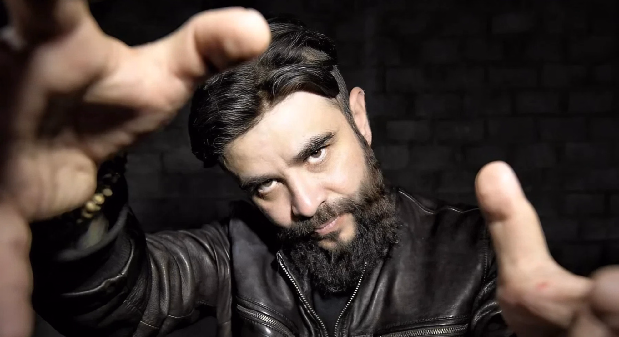 Director Carlos Ortiz Alarcón, bearded, in a black leather jacket, reaching both open hands toward the camera against a dark brick wall.
